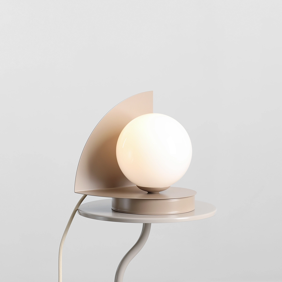 Lampa stołowa Loop Beige z jedną oprawą E14, z okrągłą, białą kulą oraz półokrągłym metalowym elementem w beżowym kolorze. Stoi na okrągłej podstawie, z widocznym kablem o jasnym oplocie. Tło jasne, minimalistyczne.