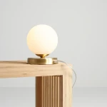 Lampa stołowa Ball D14 z mosiężną podstawą i okrągłym, matowym kloszem w kolorze białym. Ustawiona na drewnianym blacie, z widocznym kablem zasilającym. Kąt ujęcia: frontalny, podkreślający prosty design.