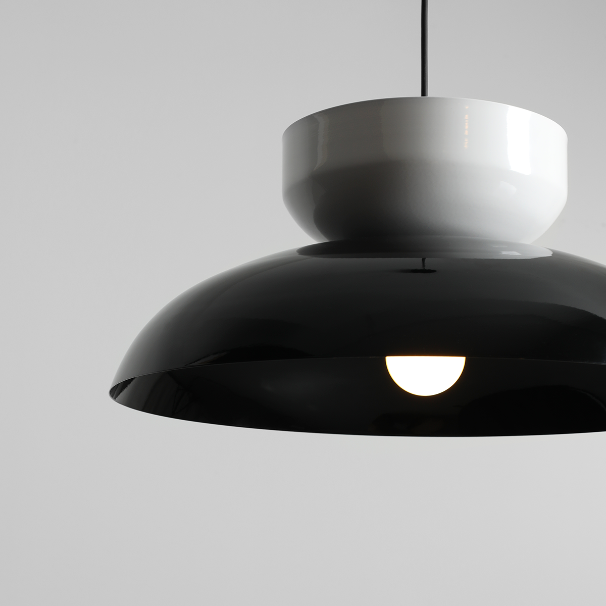 Lampa wisząca Aldo z kloszem w połyskującej czerni i białym elementem u góry. W kształcie stożka, z widocznym źródłem światła E27. Zawieszona na cienkim przewodzie, na jednolitym, jasnym tle.