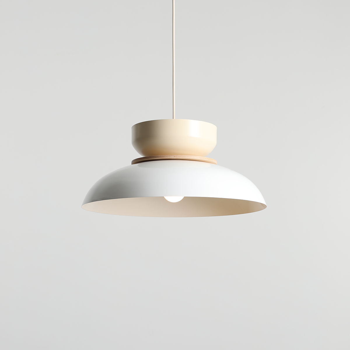 Lampa wisząca Aldo 1xE27 w kolorze białym i kremowym, z okrągłym kloszem oraz cylindrycznym elementem u góry. Zawieszona na cienkim przewodzie, prezentuje nowoczesny design. Widoczny element drewniany podkreśla estetykę lampy.