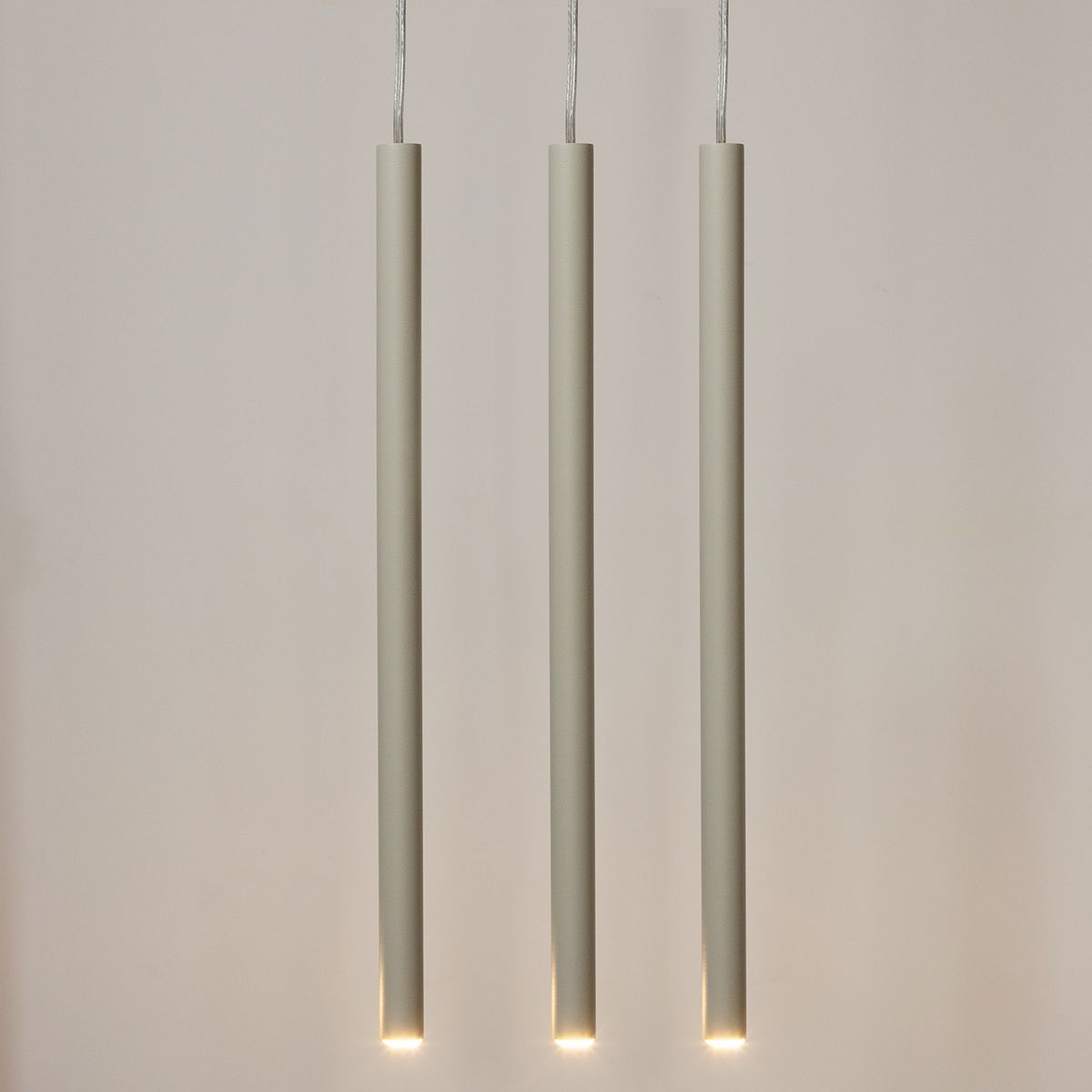 Lampa wisząca Sopel w kolorze szarym beżowym z trzema cylindrycznymi kloszami. Wisi na cienkich przewodach, każdy klosz z dolnym źródłem światła. Tło jasne, proste, podkreślające minimalistyczny design lampy.