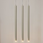 Lampa wisząca Sopel w kolorze szarym beżowym z trzema cylindrycznymi kloszami. Wisi na cienkich przewodach, każdy klosz z dolnym źródłem światła. Tło jasne, proste, podkreślające minimalistyczny design lampy.