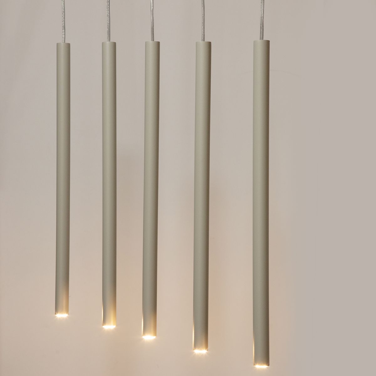 Sopel lampa wisząca w kolorze szarym beżowym, składająca się z pięciu długich, cylindrycznych elementów z delikatnym podświetleniem na końcach, zawieszona na cienkich przewodach. Idealna do nowoczesnych wnętrz.