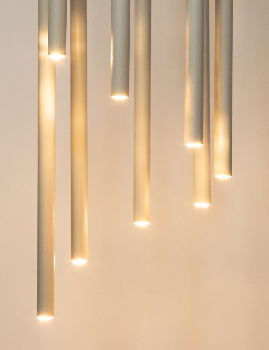 Sopel lampa wisząca w odcieniu szaro-beżowym z ośmioma rurkowymi kloszami, emitującymi światło na końcach. Produkt zawieszony w pionie, z widocznymi źródłami światła, na jasnym tle.