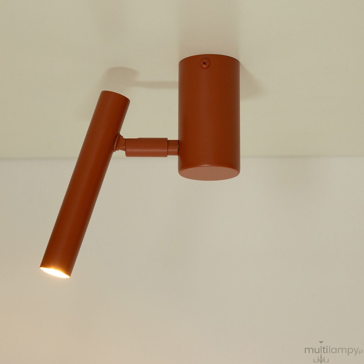 Krótka lampa sufitowa typu spot w kolorze ceglastym, z okrągłym uchwytem montażowym i prostym, wydłużonym kloszem. Ujęcie z boku, pokazujące ruchomy element lampy, skierowany w dół, na jasnym tle.
