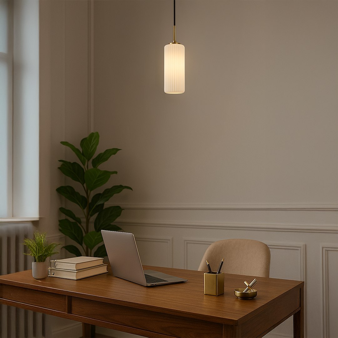 Lampa wisząca Vesta 1xE27 w kolorze złotym i białym, o cylindrycznym kształcie z żłobieniami. Powieszona na cienkim, czarnym przewodzie, w jasnym wnętrzu biurowym z drewnianym biurkiem i rośliną.