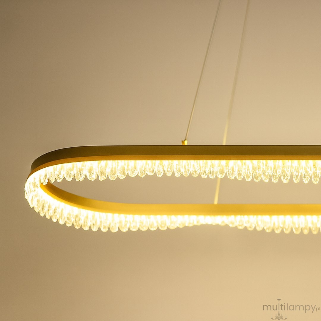 Lampa wisząca LED Preciosa 45W w złotym kolorze, o kształcie elipsy, z przezroczystymi elementami dekoracyjnymi. Ujęcie z dołu, pokazujące oświetlenie oraz detale wykończenia. Idealna do nowoczesnych wnętrz.