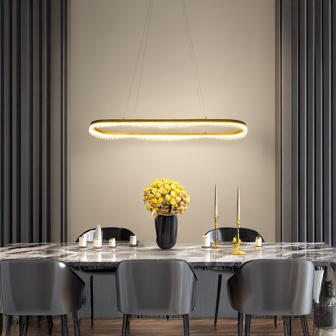 Lampa wisząca LED Preciosa 45W w kolorze złotym, o owalnym kształcie z kryształowym wykończeniem, zawieszona nad dużym stołem w eleganckiej jadalni. W tle widoczny bukiet żółtych owoców w wazonie oraz świeczniki na stole.