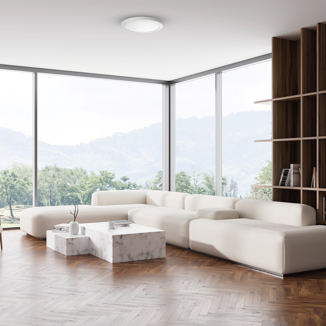 Plafon LED Evora Round D30, 24W, biały, okrągły kształt, zamontowany na suficie w jasnym salonie z dużymi oknami. W tle widoczna biała sofa i marmurowy stół.