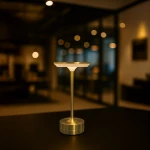 Lampa stołowa LED Tavolo 2,5W CCT w kolorze złotym, z okrągłą podstawą i płaską górą. Ujęcie z boku, produkt stoi na ciemnym stole, w tle rozmyte światła, co podkreśla elegancki design. Idealna do nowoczesnych wnętrz.