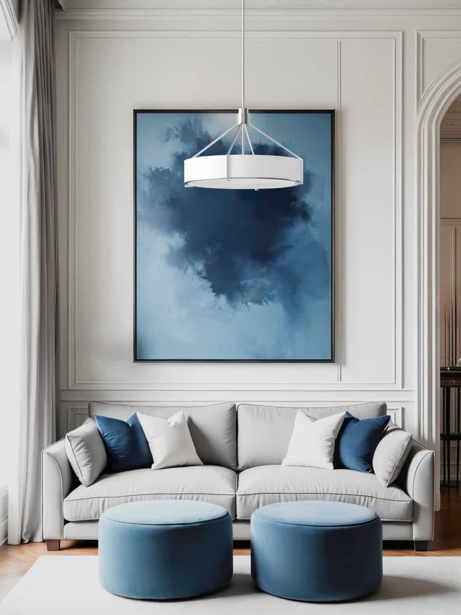 Lampa wisząca Doretto Satin Nikiel D50 w kolorze srebrnym, z trzema miejscami na żarówki E27. Okrągły klosz zawieszony w nowoczesnym wnętrzu, w tle obraz w odcieniach niebieskiego, obok szarej sofy i niebieskich pufów.