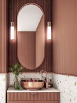 Złota lampa wisząca LED Omi I Gold 10W 3000K, umieszczona w nowoczesnej łazience. Lampa ma cylindryczny kształt z przezroczystym kloszem, zawieszona na cienkim przewodzie, obok lustra w owalnej ramie. Tło w odcieniach różu z teksturą.