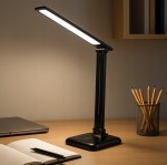 Czarna lampka biurkowa Marina LED o mocy 9W i temperaturze barwowej 4500K, z regulowanym ramieniem, stojąca na szerokiej podstawie. W tle notatnik i laptop, obok pojemnik z ołówkami.