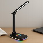 Czarna lampka biurkowa Alma LED 12W z regulowanym ramieniem, z kolorowym podświetleniem podstawy. Ujęcie z boku, na drewnianym biurku, obok otwartej książki i laptopa. Włącznik z kolorowym kółkiem na podstawie.
