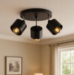 Okrągła lampa sufitowa Elza w kolorze czarnym z trzema reflektorami E27. Reflektory mają metalowe klosze w formie walca, skierowane w różne strony. Lampa zamontowana na okrągłej podstawie, w tle widoczna jasna ściana i okno z zasłonami.
