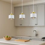 Lampa wisząca Elza z białym kloszem i złotym detalem, zawieszona na białym przewodzie. Widoczne trzy lampy w kuchennym otoczeniu, na tle szarych szafek i jasnej płyty roboczej. W tle drewniana miska z zielonymi jabłkami.