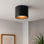 Lampa sufitowa Golda w formie czarnej tuby z złotym wnętrzem, zamontowana na suficie. Prosta, cylindryczna konstrukcja, emitująca światło, idealna do nowoczesnych wnętrz. Tło w jasnych odcieniach z roślinnym akcentem w dolnym rogu.