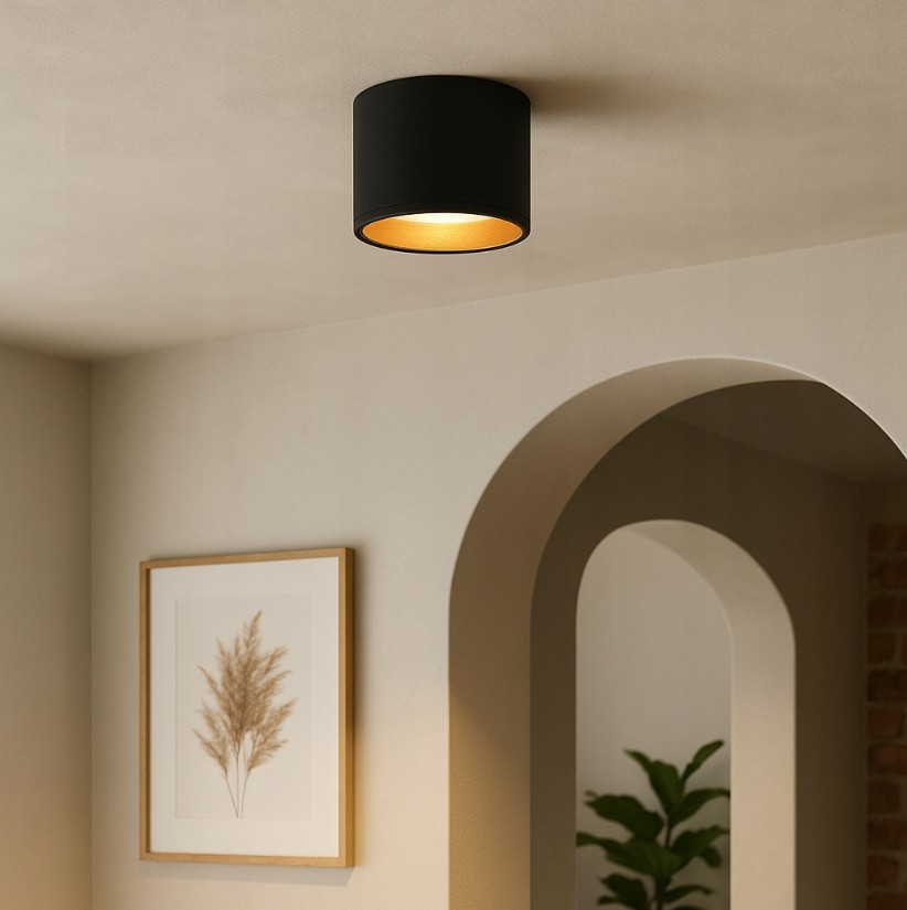 Lampa sufitowa Golda, w kształcie tuby, w kolorze czarnym z złotym wnętrzem, zamontowana na suficie. Oświetlenie o prostym, cylindrycznym designie, idealne do nowoczesnych wnętrz. Tło w stonowanej kolorystyce, z dekoracyjnym obrazem i rośliną w aranżacji.