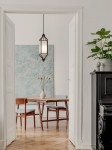 Lampa wisząca Zundo Chocco D20 z brązowym metalowym stelażem i mlecznobiałym kloszem, zawieszona w jadalni nad drewnianym stołem z niebieskimi krzesłami. W tle widoczna zielona dekoracyjna ściana oraz rośliny w doniczkach.
