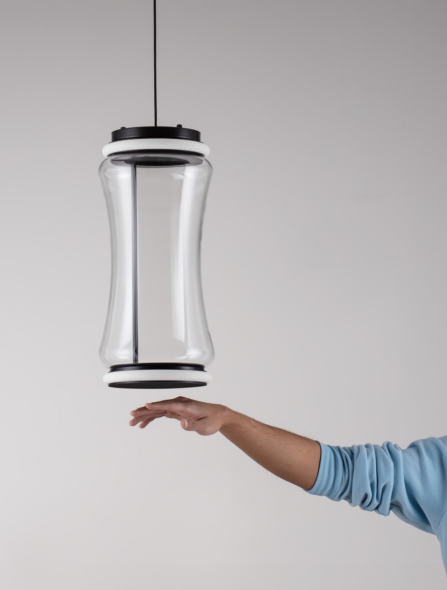 Lampa wisząca Abancay 2xLED 21W w kolorze czarnym. Posiada szklaną obudowę o falistym kształcie, zawieszona na czarnym przewodzie. Widoczna dłoń sugeruje wysokość lampy. Tło jednolite, neutralne.