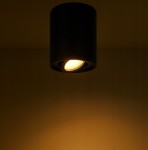 Lampa sufitowa tuba w kolorze czarnym, zawieszona na suficie, z widocznym źródłem światła w kształcie okrągłym. Kąt ujęcia z dołu, podkreślający kierunkowe oświetlenie. Tło jednolite, ciemne, co zwiększa widoczność lampy.