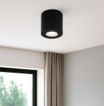 Czarna lampa sufitowa tuba Aqua IP54, zamontowana na suficie, z okrągłym źródłem światła. Widoczny jest gładki, matowy materiał oraz minimalistyczny design. Tło to jasna ściana i zasłony w odcieniu beżowym.