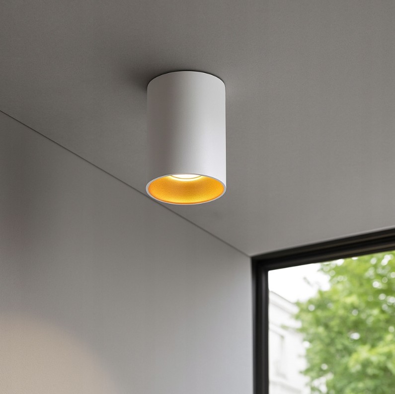 Lampa sufitowa typu tuba w kolorze białym z złotym wnętrzem, zamontowana na suficie. Okrągły kształt z widocznym źródłem światła GU10, umieszczona w rogu pomieszczenia. Tło w neutralnych barwach, z oknem w tle.