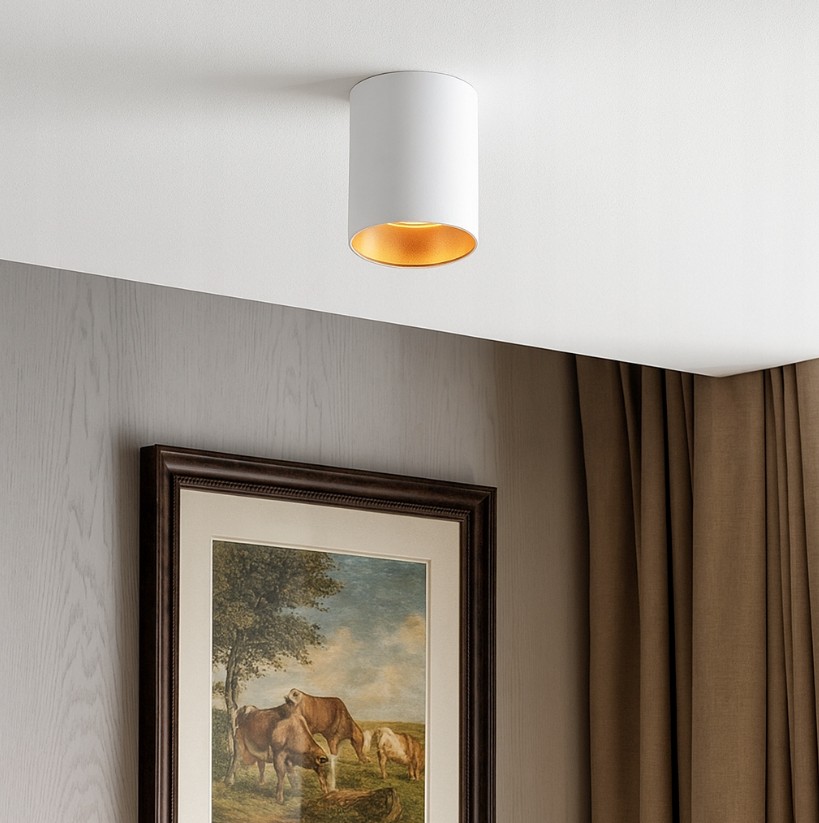Biała lampa sufitowa w kształcie tuby z złotym wnętrzem, zamontowana na suficie. Widoczna w aranżacji z obrazem na ścianie oraz zasłonami w odcieniu brązowym. Idealna do nowoczesnych wnętrz.