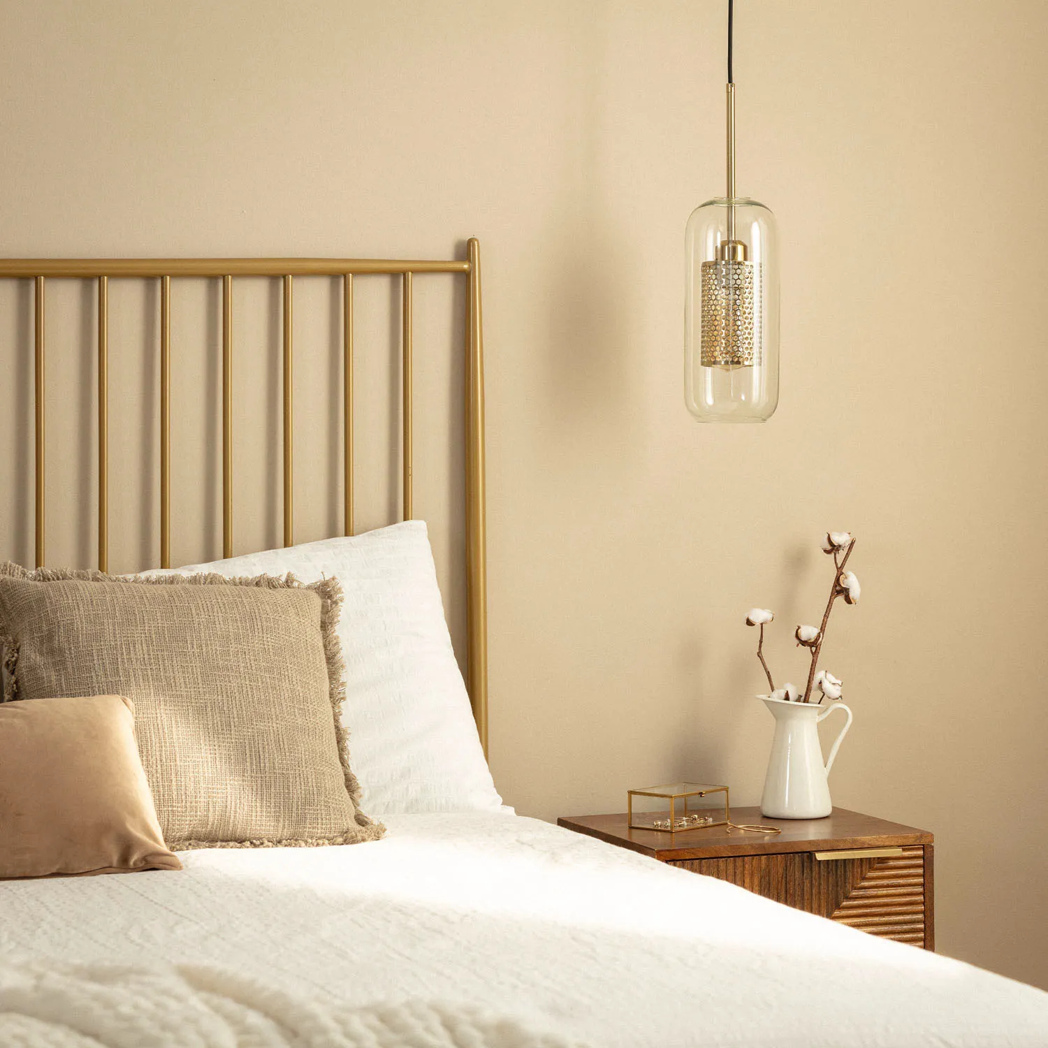 Lampa wisząca Nobbile z jedną oprawą E27 w złotym kolorze, zawieszona nad łóżkiem. Szklany klosz z dekoracyjnym wkładem, na tle jasnej ściany. W aranżacji z poduszkami w beżowych odcieniach i drewnianą szafką nocną.