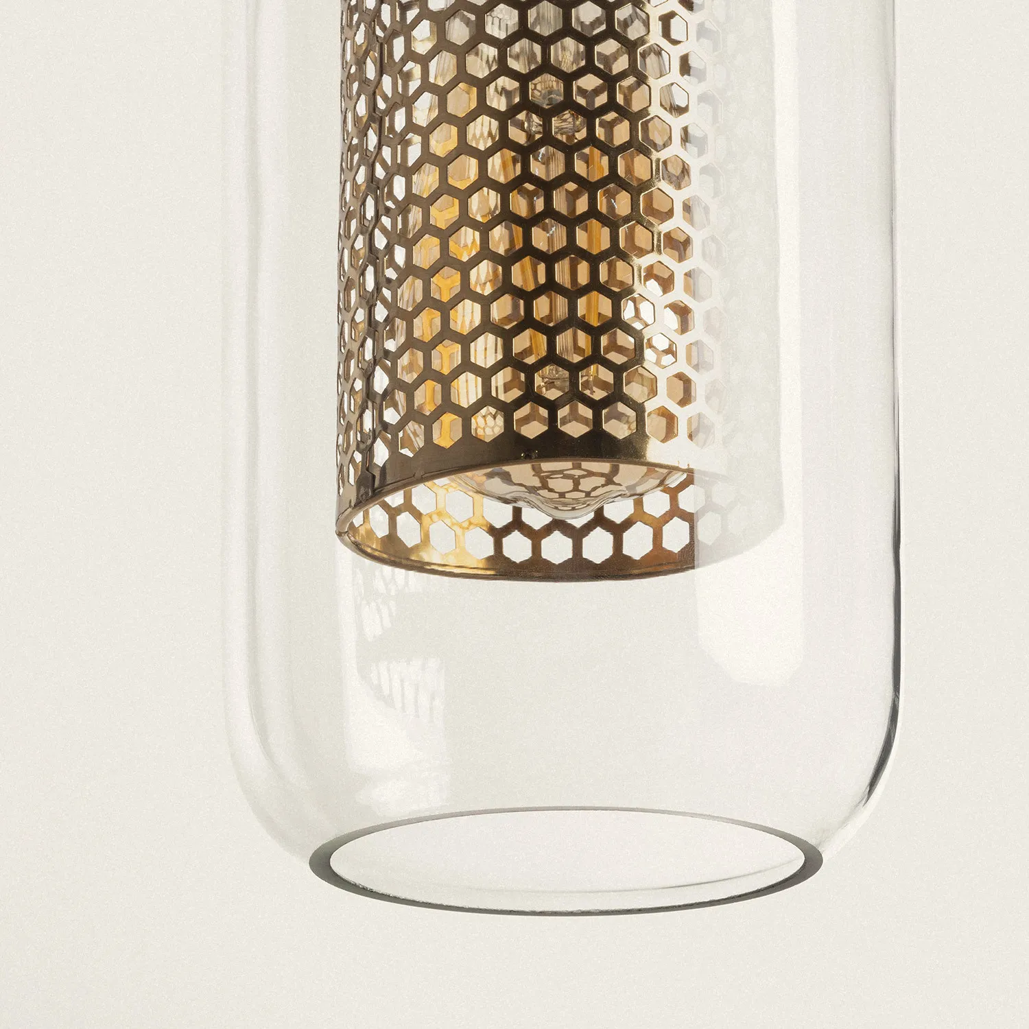 Lampa wisząca Nobbile z oprawą w złotym kolorze, z metalowym kloszem o wzorze heksagonalnym. Ujęcie z dołu, ukazujące przezroczysty szklany element. Przeznaczenie na żarówki E27. Tło jasne i jednolite.