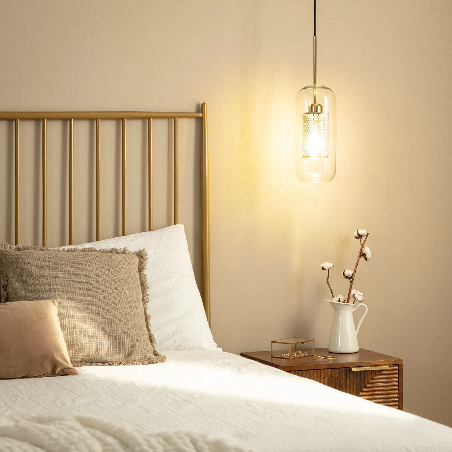 Złota lampa wisząca Nobbile z kloszem w kształcie prostokątnym, zawieszona nad łóżkiem. W tle widoczna zagłówka łóżka z metalowymi prętami, białe pościele i poduszki, stolik nocny z białym dzbankiem i kwiatami.