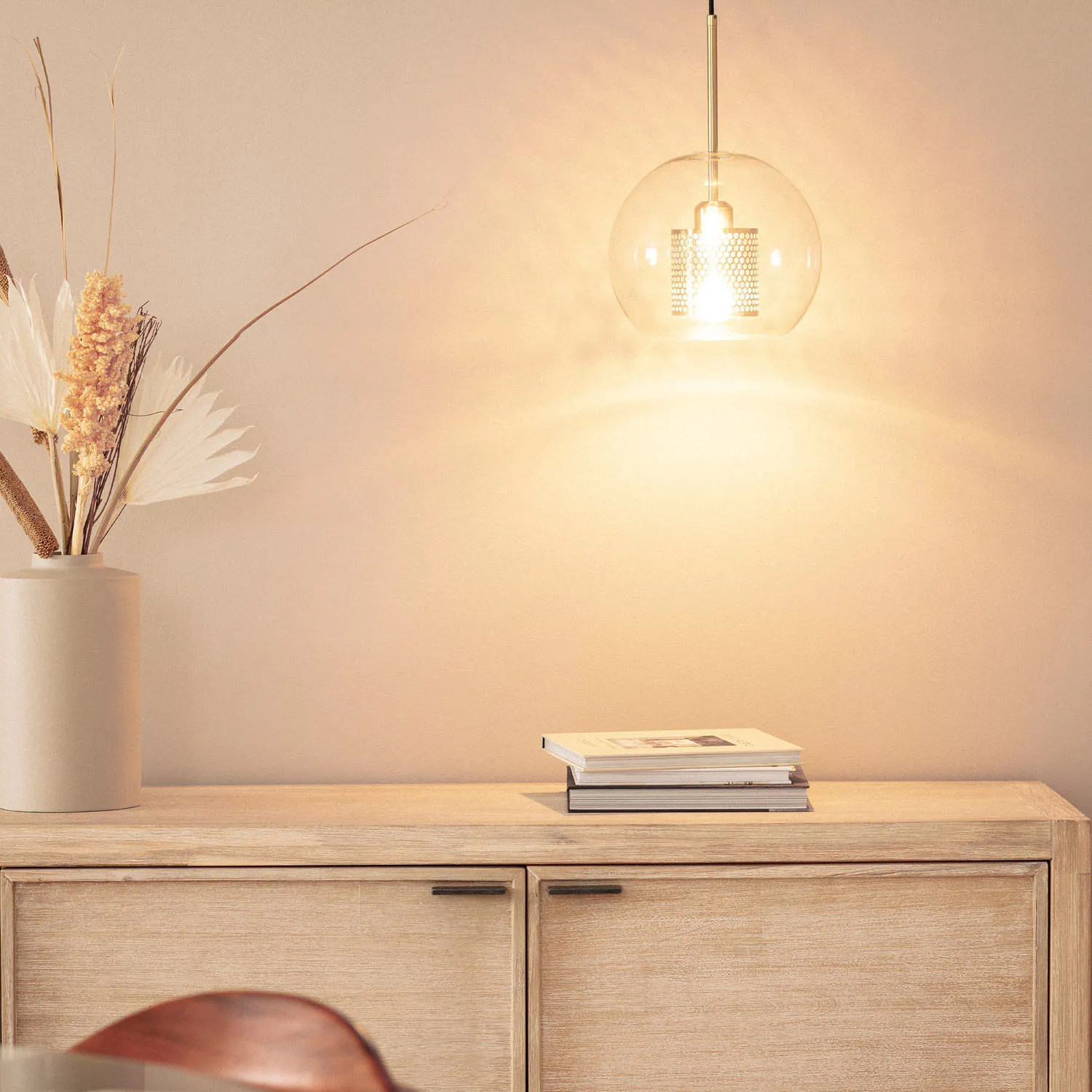 Lampa wisząca Nobbile 2, z pojedynczym źródłem światła E27, w złotym kolorze. Szklany klosz o okrągłym kształcie, zawieszony na metalowym stelażu. Tło w spokojnym odcieniu, obok wazon z dekoracyjnymi roślinami i stos książek.