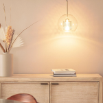 Lampa wisząca Nobbile 2, z pojedynczym źródłem światła E27, w złotym kolorze. Szklany klosz o okrągłym kształcie, zawieszony na metalowym stelażu. Tło w spokojnym odcieniu, obok wazon z dekoracyjnymi roślinami i stos książek.