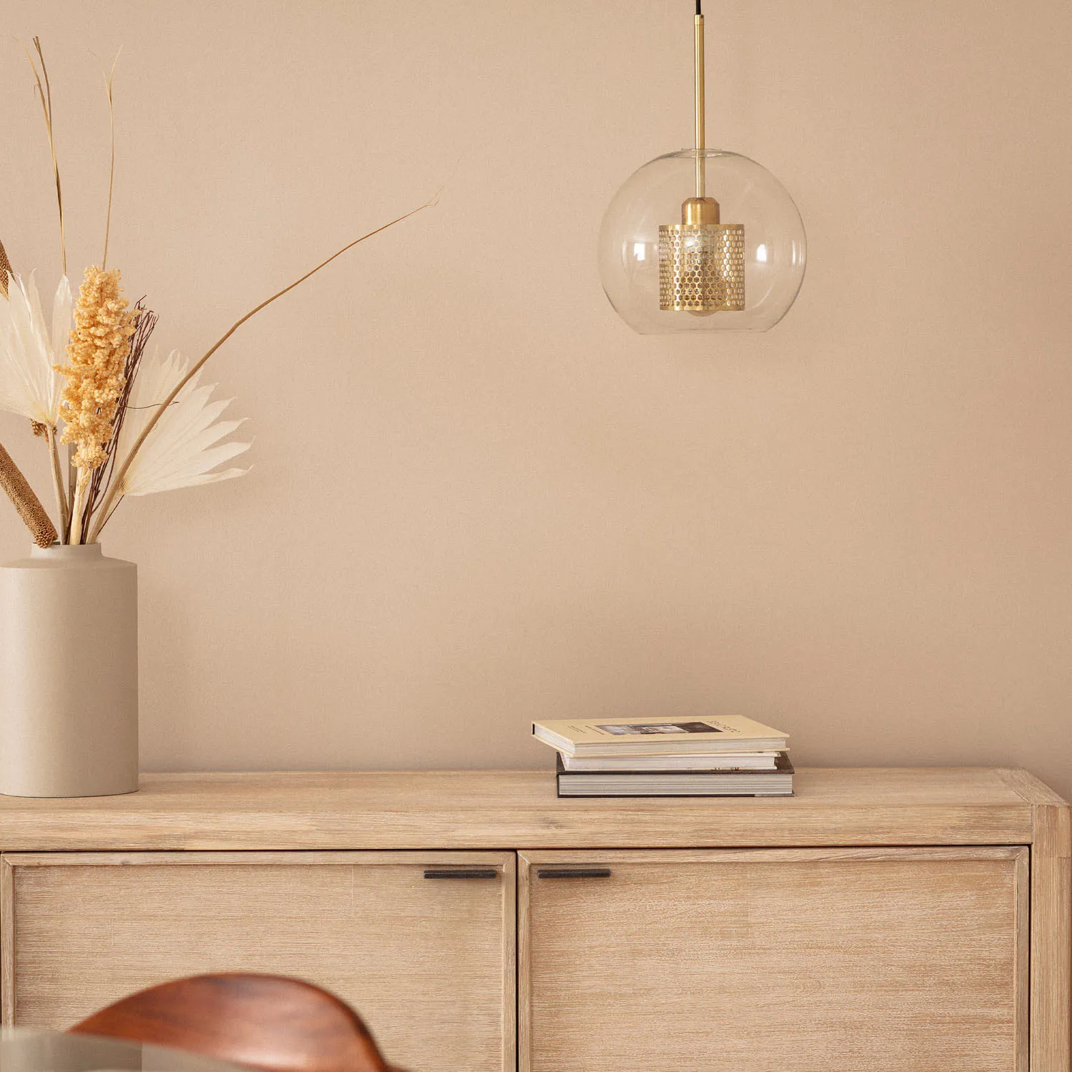 Lampa wisząca Nobbile 2, z jednolitym, przezroczystym kloszem i złotym detalem, zawieszona nad drewnianym meblem. W tle wazon z suszonymi roślinami oraz stos książek. Tło w neutralnym odcieniu beżu.