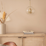 Lampa wisząca Nobbile 2, z jednolitym, przezroczystym kloszem i złotym detalem, zawieszona nad drewnianym meblem. W tle wazon z suszonymi roślinami oraz stos książek. Tło w neutralnym odcieniu beżu.