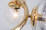 Lampa wisząca Amaro w kolorze złotym, z pięcioma oprawami E14. Ujęcie zbliżeniowe na jedną z kulistych szklanych kloszy, w której widoczna jest żarówka. Elementy metalowe mają błyszczącą fakturę, a klosz jest transparentny.