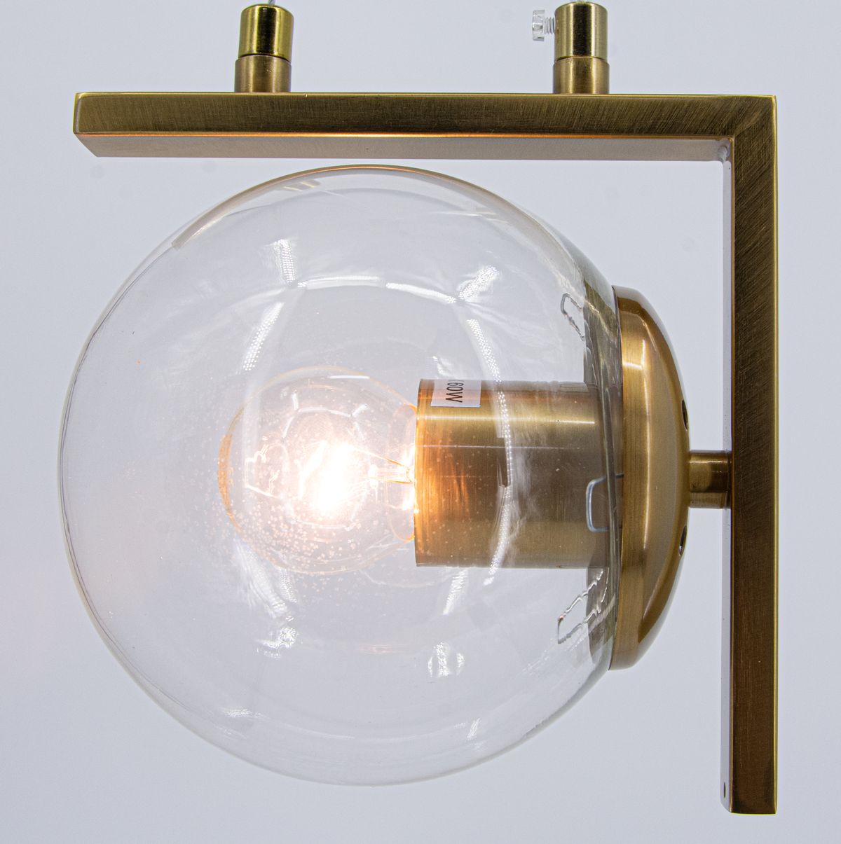Lampa wisząca Brava z jedną oprawą E27, wykonana z mosiądzu. Szklana kula osłania żarówkę, zamocowana na prostokątnej, mosiężnej podstawie. Ujęcie z boku, z widocznym źródłem światła w środku.