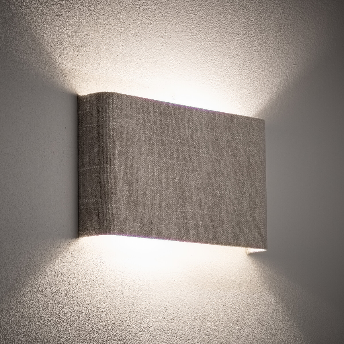 Kinkiet Santana B w kolorze kremowym, z tkaninowym abażurem, montowany na ścianie. Oświetlenie emituje delikatne światło, tworząc subtelne cienie na ścianie. Prosty, prostokątny kształt, zamontowany poziomo.