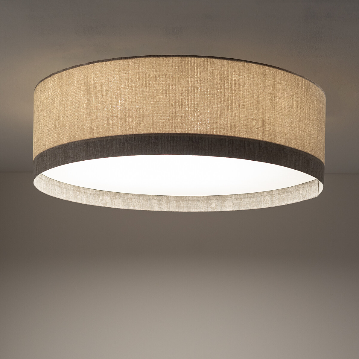 Lampa sufitowa Cedro M w kolorze beżowym, o okrągłym kształcie z materiałowym abażurem. Widoczny szary pasek na krawędzi. Ujęcie z dołu, ukazujące gładką, białą powierzchnię wewnętrzną. Przeznaczona na żarówkę E27.
