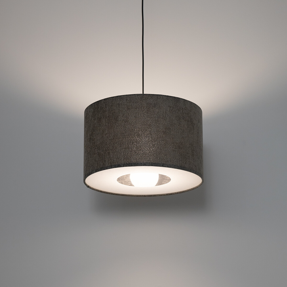 Lampa wisząca Lago S w kolorze szarym z kloszem w kształcie cylindrycznym, zawieszona na cienkim przewodzie. Wnętrze klosza białe, widoczna żarówka E27, emitująca ciepłe światło, na tle gładkiej, jasnej ściany.