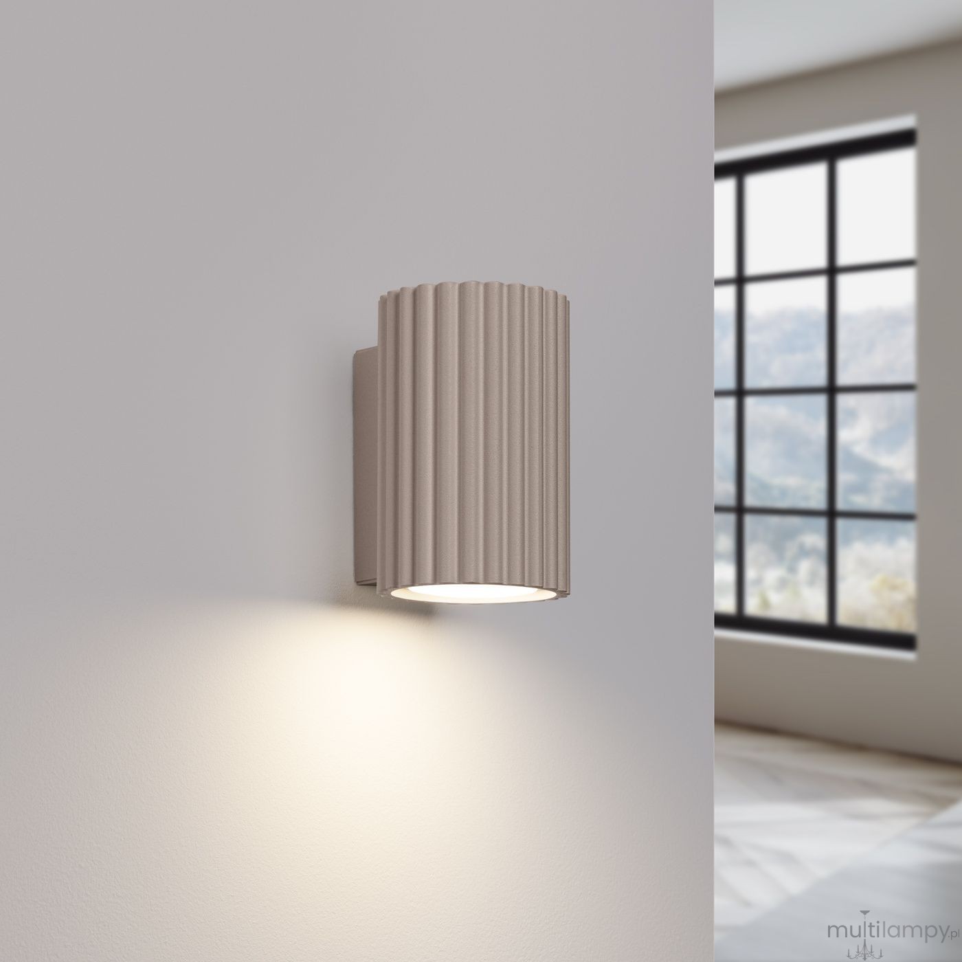 Kinkiet Karbon 10 w kolorze taupe, o cylindrycznym kształcie z rowkowaną powierzchnią. Zainstalowany na ścianie, emitujący ciepłe światło. Tło z jasną ścianą oraz oknem w tle, co podkreśla elegancki design lampy.