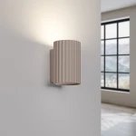 Kinkiet Karbon 10 w kolorze taupe z jedną oprawą GU10, o cylindrycznym kształcie z rowkowaną fakturą. Zainstalowany na ścianie, emitujący światło ku górze, w tle widoczne okno z widokiem na zewnątrz.