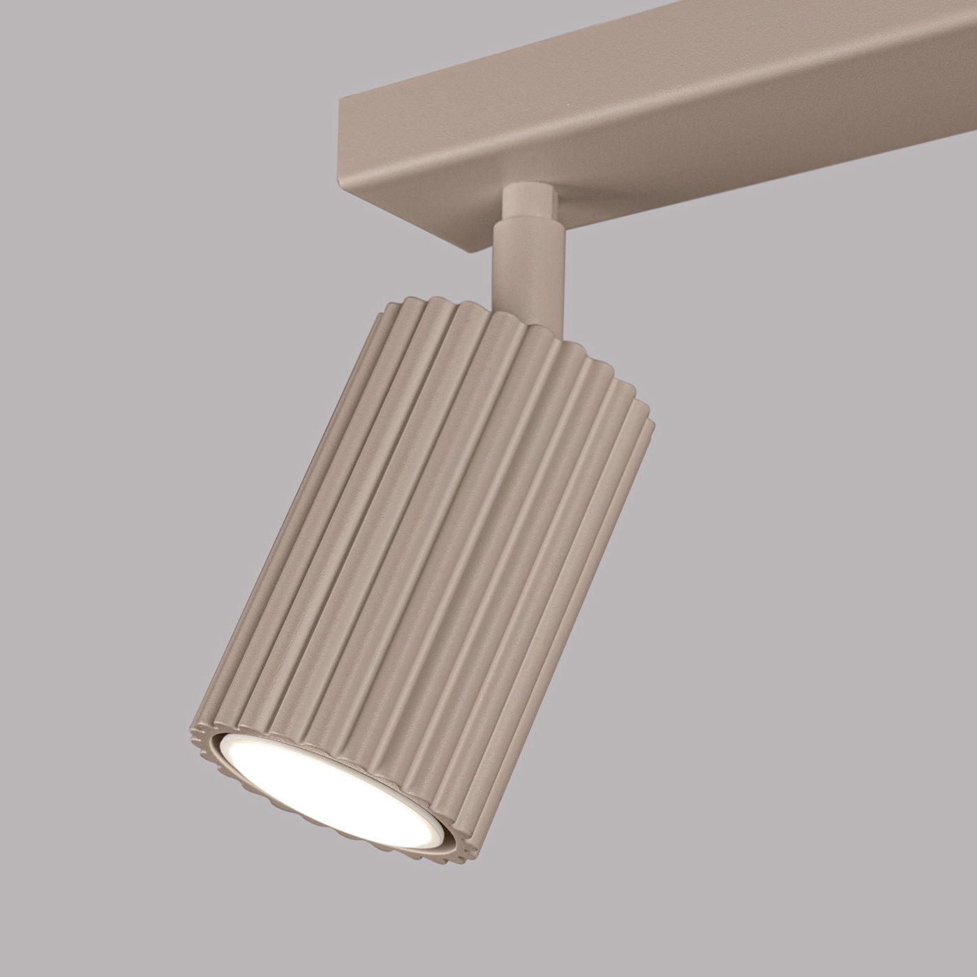 Lampa sufitowa Karbon w kolorze taupe, z dwoma spotami GU10. Kształt cylindryczny z widocznymi rowkami na powierzchni, zamontowana na prostokątnej podstawie. Źródło światła skierowane w dół, z jasną żarówką. Tło jednolite, szare.