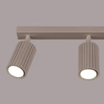 Lampa sufitowa Karbon w kolorze taupe z trzema reflektorami GU10. Ujęcie z przodu, pokazujące dwa cylindryczne klosze z żebrowaną fakturą, zamocowane na prostokątnej podstawie. Klosze emitują jasne światło, tło w jednolitym, stonowanym odcieniu.