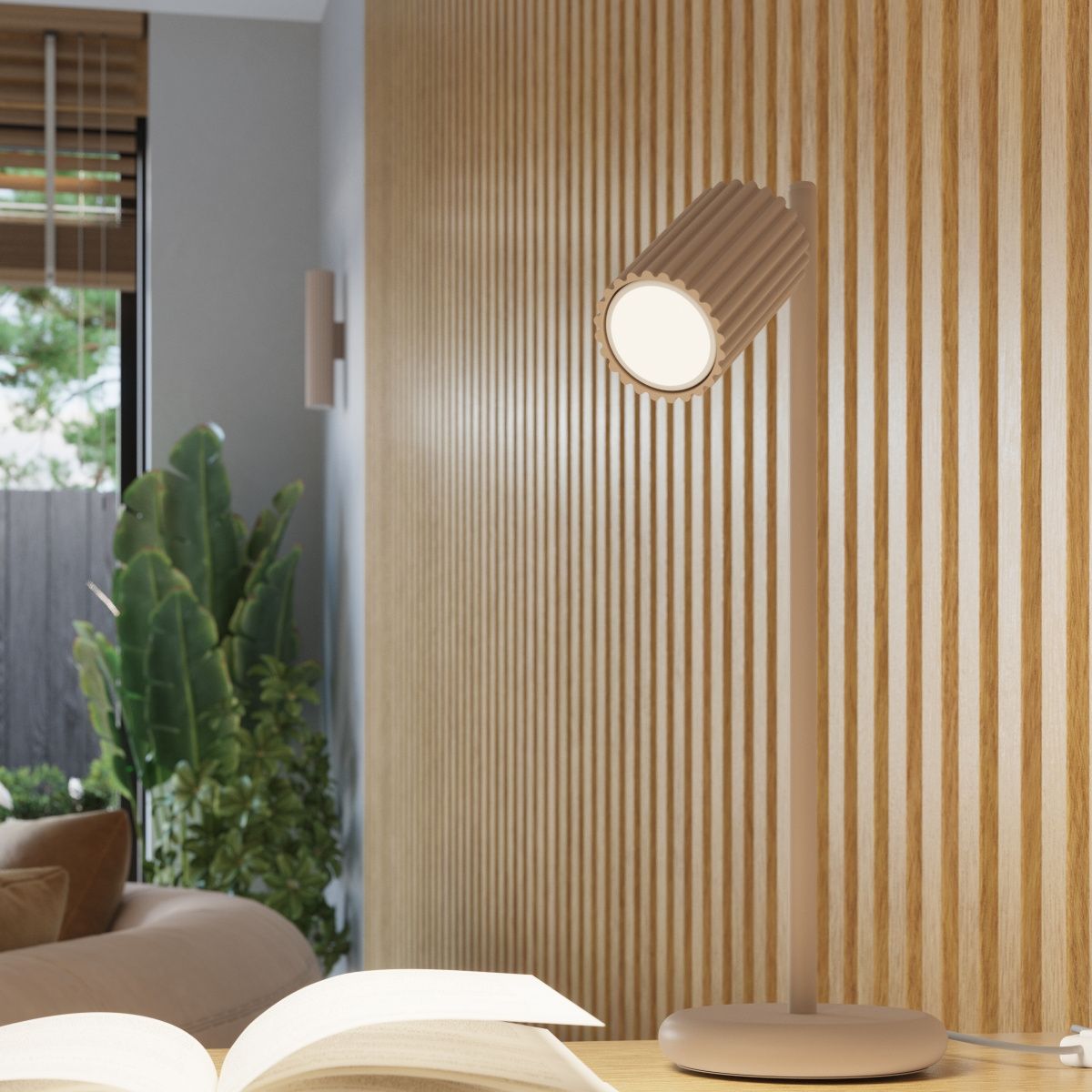 Lampka stołowa Karbon 1xGU10 w kolorze taupe z cylindrycznym kloszem o żłobieniach. Stojąca na okrągłej podstawie, skierowana w bok. W tle widoczna drewniana ściana i rośliny, co tworzy przytulną atmosferę.