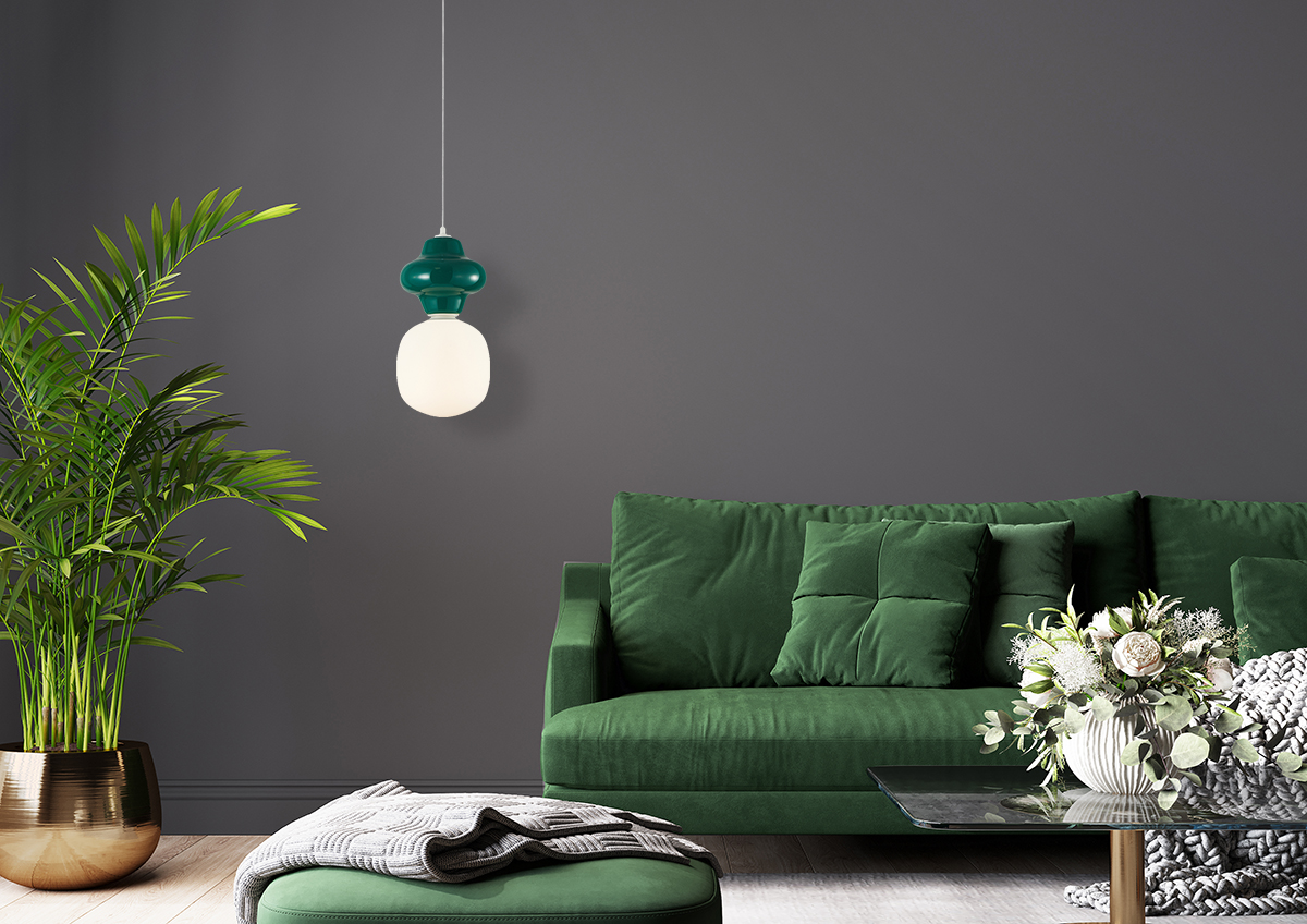 Lampa wisząca Latera D15 w kolorze zielonym z oprawą z tworzywa. Posiada okrągły klosz z białego szkła, zawieszona na cienkim przewodzie. Umieszczona nad zieloną sofą w nowoczesnym wnętrzu z roślinnością i dekoracyjnym stolikiem.