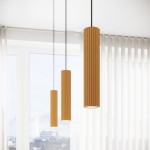 Lampa wisząca Karbon 1 z jedną oprawą GU10 w złotym kolorze. Posiada cylindryczny kształt z rowkowaną fakturą, zawieszona na czarnym kablu. Wisi w grupie trzech lamp o różnych wysokościach, na tle jasnego wnętrza z dużymi oknami.