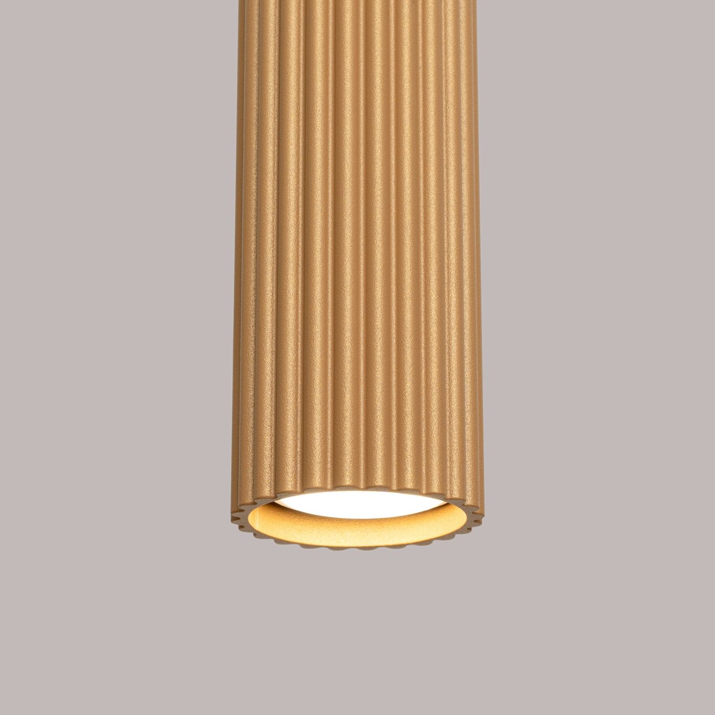 Lampa wisząca Karbon w kolorze złotym, z cylindrycznym kloszem o fakturze żłobionej. Ujęcie zbliżeniowe pokazuje górną część lampy oraz oprawę na żarówkę GU10. Tło w odcieniu szaro-beżowym, minimalistyczne.