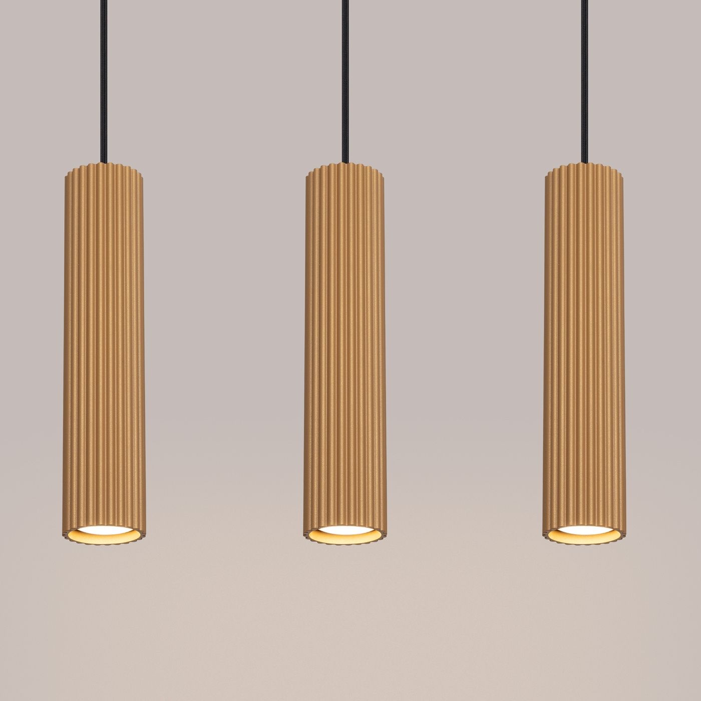 Lampa wisząca Karbon 3 w kolorze złotym, z trzema cylindrycznymi kloszami z rowkowanej powierzchni, zawieszona na czarnych przewodach. Klosze mają otwarte dolne części, co umożliwia montaż żarówek GU10. Tło w jednolitym, jasnym odcieniu.