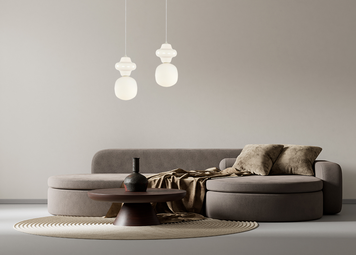 Kremowa lampa wisząca Latera D15 z jednym gniazdem E14, o zaokrąglonym kształcie, zawieszona na cienkim przewodzie. W tle nowoczesny, szary narożnik oraz okrągły stół z ciemnego drewna, w aranżacji na jasnym tle.
